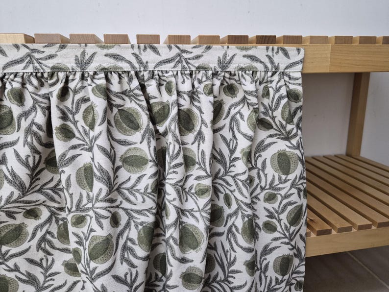 May include: A close-up of a fabric curtain with a botanical pattern. The curtain features a white background with a repeating design of green pomegranates and gray leaves. The curtain is attached to a wooden shelf.