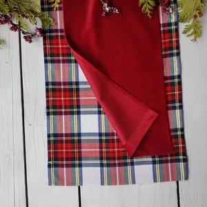 May include: A red and plaid table runner. The runner features a solid red top layer and a plaid bottom layer with red, green, white, and blue accents. The runner is displayed on a white wooden surface, with greenery and berries in the background.