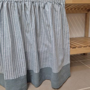May include: A light blue and white striped fabric curtain with a solid blue-gray border. The curtain is hanging from a wooden shelf, creating a decorative and functional element.