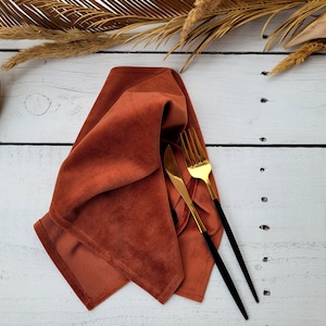 Puede incluir: Una servilleta de terciopelo color óxido doblada sobre una superficie de madera blanca con un tenedor dorado y un cuchillo con mango negro.