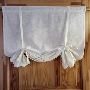 Könnte beinhalten: Ein weißer Fensterbehang aus Leinen mit Bindebändern. Der Behang ist am unteren Rand gerafft und mit Schleifen auf jeder Seite gebunden. Der Stoff ist ein leichtes, strukturiertes Material, und der Behang hängt an einer Holztür.