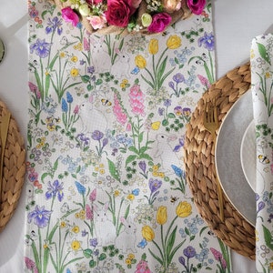 Może przedstawiać: Biały bieżnik stołowy z kwiatowym wzorem przedstawiającym króliki, kwiaty i liście. Bieżnik leży na białym stole z dwoma nakryciami, każde z plecionym podkładką, białym talerzem i złotym widelcem.