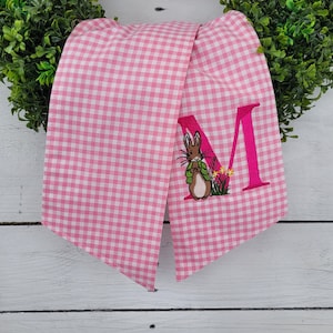 May include: Pink and white gingham fabric scarf with an embroidered letter "M" and a cartoon rabbit wearing a green jacket. The scarf is draped over a green wreath, creating a spring or Easter theme.