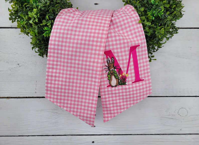 May include: Pink and white gingham fabric scarf with an embroidered letter "M" and a cartoon rabbit wearing a green jacket. The scarf is draped over a green wreath, creating a spring or Easter theme.