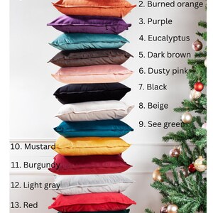 Op de afbeelding: Een stapel van 15 fluwelen kussens in verschillende kleuren, waaronder marineblauw, gebrand oranje, paars, eucalyptus, donkerbruin, poederroze, zwart, beige, mosterd, bordeauxrood, lichtgrijs, rood, groen en donkergrijs. De kussens zijn gestapeld van boven naar beneden, met de kleur van elk kussen ernaast vermeld. De afbeelding bevat ook een kleine kerstboom met kerstballen.