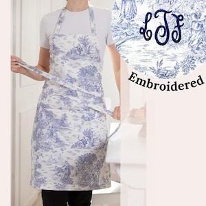 Könnte beinhalten: Eine weiße Schürze mit einem blauen und weißen Toile-Muster. Die Schürze hat eine Tasche und wird in der Taille gebunden. Die Schürze ist mit den Initialen "L.J.F." bestickt.