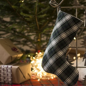 Könnte beinhalten: Ein grüner und weißer karierter Weihnachtssocken hängt an einem Metall-Sockenhalter mit Engel- und Baumschmuck. Der Socken ist mit Geschenken gefüllt und von einem Weihnachtsbaum und Lichtern umgeben.