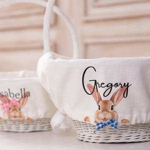 May include: Two white wicker Easter baskets with fabric liners. One basket has a bunny with a pink bow and the name "Isabella" printed on it. The other basket has a bunny with a blue bow and the name "Gregory" printed on it.
