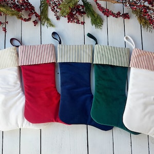 May include: Five Christmas stockings with different colored velvet fabric and white cotton cuff with a striped pattern. The stockings are arranged in a row on a white wooden surface.