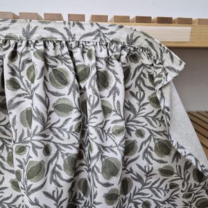 May include: A white fabric with a repeating pattern of green pomegranates and gray leaves. The fabric is gathered at the top, creating soft folds. The design is printed on a light-colored background.
