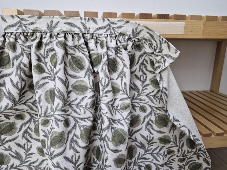 May include: A white fabric with a repeating pattern of green pomegranates and gray leaves. The fabric is gathered at the top, creating soft folds. The design is printed on a light-colored background.