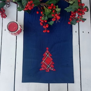 May include: A blue table runner with a red and black plaid Christmas tree design. The runner is on a white wooden surface with red berries and green leaves.