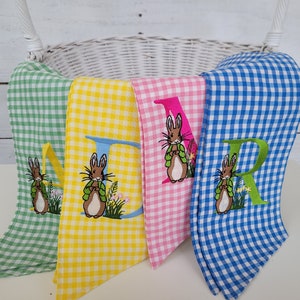 May include: Four gingham fabric Easter decorations in green, yellow, pink, and blue. Each has a large embroidered letter and a bunny with a green vest. The decorations are in front of a white wicker basket.