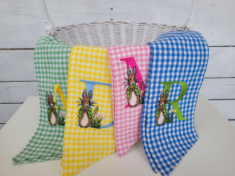 May include: Four gingham fabric Easter decorations in green, yellow, pink, and blue. Each has a large embroidered letter and a bunny with a green vest. The decorations are in front of a white wicker basket.