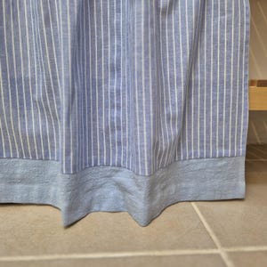May include: A blue and white striped curtain with a solid blue bottom hem. The curtain is made of a lightweight fabric and hangs in soft folds. The stripes are vertical and evenly spaced.