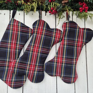 Op de afbeelding: Drie kerstsokken in een klassiek ruitpatroon van rood, zwart, geel en blauw. Twee zijn botvormig, één heeft de klassieke sokvorm. Ze hangen tegen een witte houten achtergrond met kerstgroen.