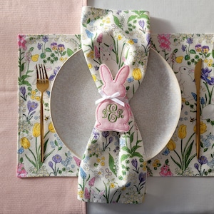 Peut inclure: Un anneau de serviette en forme de lapin rose avec les initiales "J.G.R." brodées dessus. L'anneau de serviette est placé sur une serviette blanche à motif floral. La serviette est sur une assiette blanche avec un dessous de plat à motif floral.