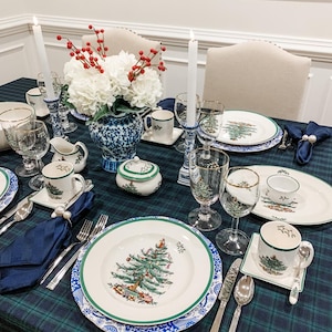May include: A festive Christmas table setting with a plaid tablecloth. The table is set with white plates featuring a Christmas tree design, complemented by matching cups, saucers, and glassware. A blue and white vase holds white flowers and red berries.