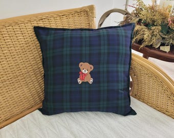 May include: A blue and green plaid throw pillow with a brown teddy bear embroidered on it. The teddy bear is wearing a red bow.