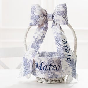 May include: White wicker basket with a blue and white patterned fabric bow and lining. The fabric has a toile pattern and the name "Mateo" is embroidered on the bow and lining.