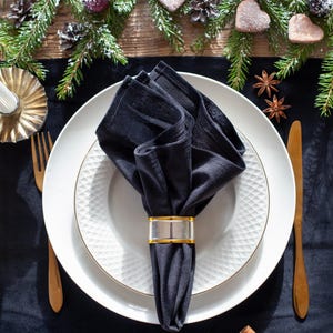 May include: A festive table setting with a black napkin held by a gold and clear napkin ring, placed on a white plate with a gold rim. The table is decorated with pine branches, ornaments, and cinnamon sticks, creating a holiday theme.