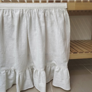 May include: A white linen skirt with a ruffled hem, hanging from a wooden shelf. The skirt has a textured appearance and a simple, elegant design. The shelf is made of light-coloured wood.