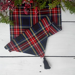 May include: A red, black, blue and yellow tartan table runner with a black tassel. The runner is folded in half and has a white background.