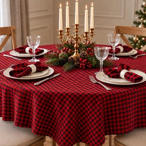 May include: A round table covered with a red and black buffalo check tablecloth, set with white plates, clear glasses, and napkins in white rings. A gold candelabra with five white candles sits in the center with pine and berry decor.
