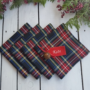May include: Four red, black, and green plaid napkins with a red ribbon and a small bell. A red tag with the name "Kate" is attached to the ribbon.