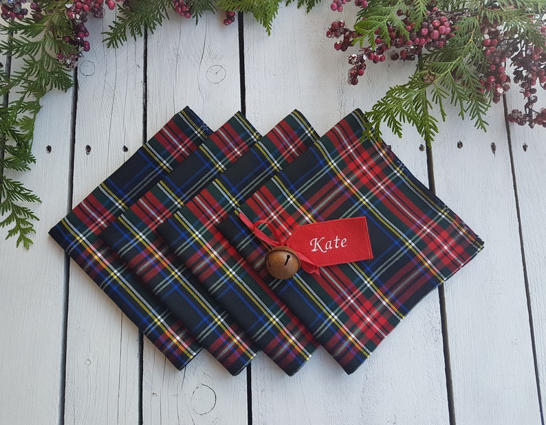 May include: Four red, black, and green plaid napkins with a red ribbon and a small bell. A red tag with the name "Kate" is attached to the ribbon.