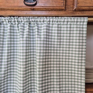 May include: A sage green and white checkered kitchen curtain hangs below a wooden cabinet. The curtain is gathered at the top and has a simple, classic design. A stack of white plates is visible to the right.