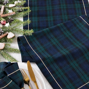 May include: A dark green and navy blue plaid table runner and matching napkin featuring a gold wavy stitched border. The napkin is held by a silver metal ring and placed on a white plate next to a gold fork and knife set on a white surface.