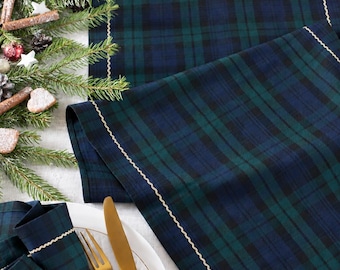 May include: A dark green and navy blue plaid table runner and matching napkin featuring a gold wavy stitched border. The napkin is held by a silver metal ring and placed on a white plate next to a gold fork and knife set on a white surface.