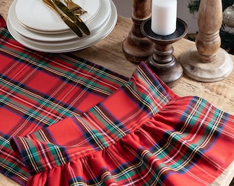 Pode incluir: Um caminho de mesa xadrez vermelho com borda franzida, colocado sobre uma mesa de madeira. Velas brancas em castiçais de madeira, talheres dourados e pratos brancos com bordas douradas completam a decoração.
