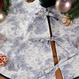 May include: A white and blue toile Christmas tree skirt with a dark blue trim. The skirt is decorated with a classic pastoral scene and tied with matching bows. Presents and ornaments surround the tree.