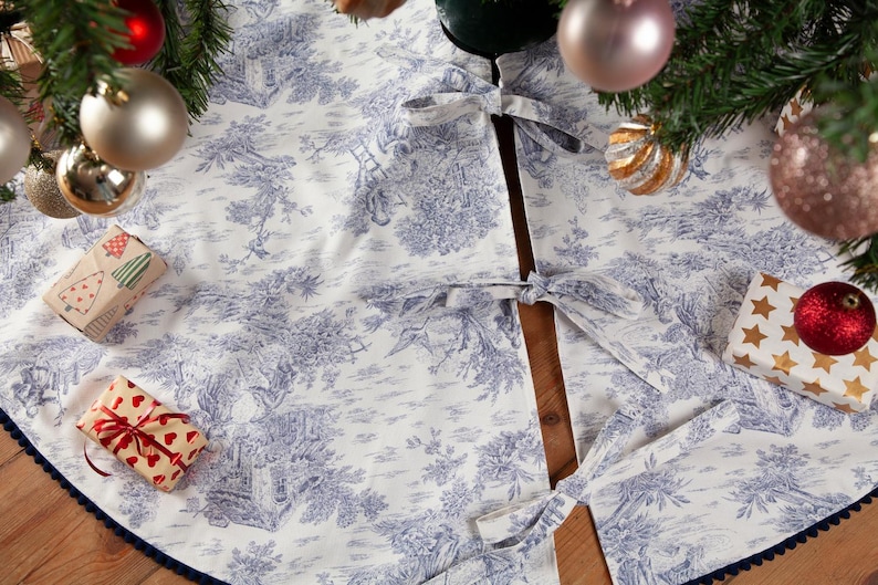 May include: A white and blue toile Christmas tree skirt with a dark blue trim. The skirt is decorated with a classic pastoral scene and tied with matching bows. Presents and ornaments surround the tree.