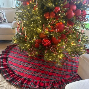May include: A decorated Christmas tree with red ornaments, berries, and lights. A red and green plaid tree skirt with a ruffled edge sits at the base of the tree. The tree is in a living room setting.