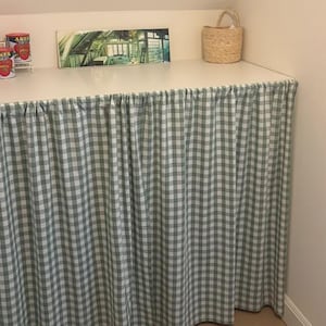 Peut inclure: Un rideau à carreaux verts et blancs recouvre le devant d'un comptoir blanc. Un petit panier tressé avec une poignée est posé sur le comptoir. Des conserves et une image encadrée sont également sur le comptoir.