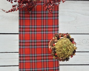 May include: Red and black plaid table runner on a white wooden surface. A small decorative arrangement with moss, pine cones, and berries is on the table runner.