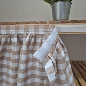 Può includere: Un'orlatura in vichy marrone e bianco con una chiusura a strappo bianca. L'orlatura è appesa a uno scaffale in legno.