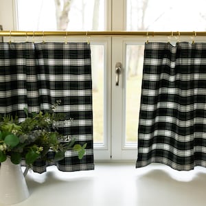 May include: A pair of plaid kitchen curtains in black, white, and green hang from a gold-colored rod above a window. A white vase with greenery sits on the windowsill. The curtains have a classic, farmhouse style.