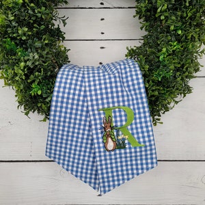 May include: A blue and white gingham patterned towel with an embroidered Peter Rabbit design and the letter "R". The towel is draped over a green wreath, creating a decorative Easter theme.
