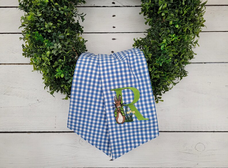 May include: A blue and white gingham patterned towel with an embroidered Peter Rabbit design and the letter "R". The towel is draped over a green wreath, creating a decorative Easter theme.