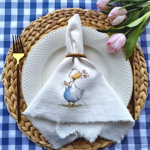 May include: A white linen napkin with a watercolor illustration of a blue duck holding flowers, set on a white plate with a gold rim. The plate rests on a woven placemat with a gold fork. Pink tulips and a blue and white checkered tablecloth complete the setting.