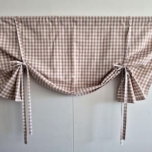 Peut inclure: Une cantonnière à carreaux marron et blanc avec des attaches. Le tissu est drapé et noué de chaque côté, créant un traitement de fenêtre décoratif. La cantonnière est faite d'un matériau doux et tissé.