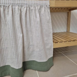 Puede incluir: Una cortina de tela a rayas de color claro con un borde verde cuelga frente a una estantería de madera. La cortina tiene rayas verticales y está fruncida en la parte superior. La estantería de madera tiene un acabado natural.