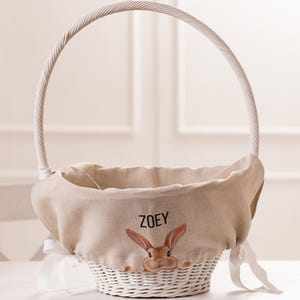 Könnte beinhalten: Weißer Weidenkorb für Ostern mit einer beigefarbenen Leinen-Innenauskleidung. Die Auskleidung zeigt ein braunes Kaninchen, das herausguckt, und den Namen "ZOEY" in brauner Farbe.