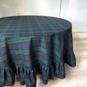 May include: A round tablecloth featuring a dark green and navy blue plaid pattern. The fabric drapes over a circular surface and is finished with a gathered ruffle trim along the bottom edge, creating a layered look with consistent textile texture.