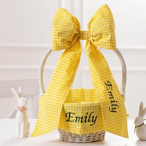 May include: A white wicker Easter basket with a yellow and white gingham bow and liner. The basket is personalized with the name "Emily" in green embroidery.
