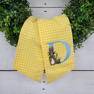 May include: Yellow and white gingham fabric with a blue embroidered letter "D" and a bunny wearing a green jacket. The bunny is standing in a patch of flowers. The fabric is draped over a green wreath.
